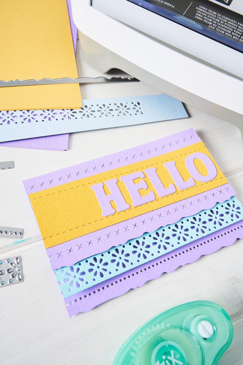 May include: A handmade greeting card with the word "HELLO" in large, light purple letters. The card features layers of yellow, light purple, and blue paper with decorative cut-out edges. A craft machine and glue are visible.