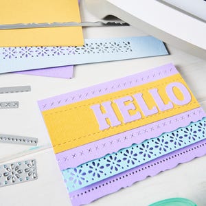 May include: A handmade greeting card with the word "HELLO" in lavender letters. The card features layers of yellow, lavender, and light blue paper with decorative cut edges and stitching details. Craft supplies are scattered around.