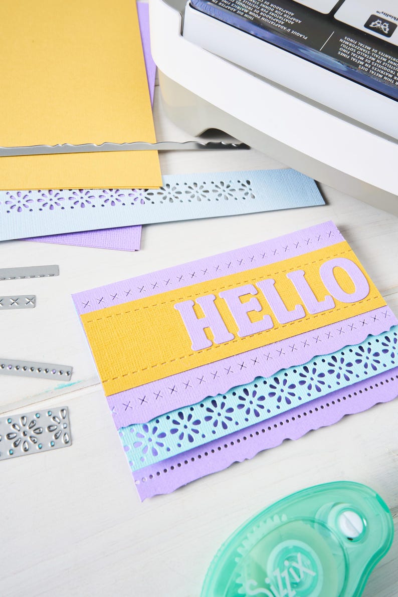 May include: A handmade greeting card with the word "HELLO" in lavender letters. The card features layers of yellow, lavender, and light blue paper with decorative cut edges and stitching details. Craft supplies are scattered around.