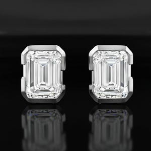 Op de afbeelding: Een paar sprankelende, rechthoekig geslepen diamanten oorstekers. Elke oorbel heeft een grote, heldere diamant met smaragdgroene slijpvorm, gezet in een zilveren of platina bezel setting. De oorbellen worden gepresenteerd tegen een zwarte achtergrond.