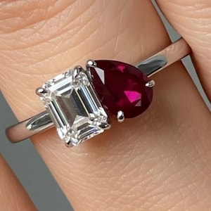 May include: A silver ring with an emerald-cut diamond and a pear-shaped ruby. The gemstones are set together, showcasing a clear diamond and a deep red ruby. The ring has a polished silver band.
