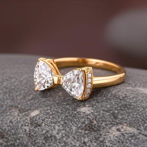 May include: A gold ring with a bow-tie design. The ring features two triangular-cut, clear gemstones at the center, with smaller stones set along the sides. The band is a polished gold color, and the ring is set on a gray stone.