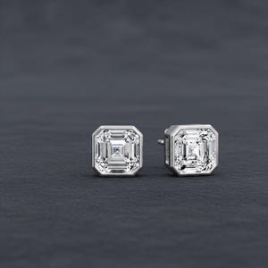 Op de afbeelding: Een paar vierkant geslepen diamanten oorstekers. De diamanten zijn gezet in een zetting met zilveren of witgouden rand. De oorbellen worden gepresenteerd op een donkergrijze ondergrond.