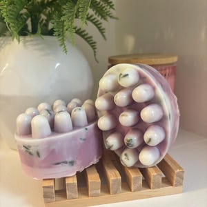 May include: Two lavender massage soaps with white, rounded protrusions, on a small wooden rack. The soaps have a marbled appearance with green flecks. A white vase with green foliage is in the background.