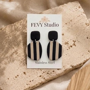 May include: A pair of black and beige striped earrings from FEVY Studio. The earrings have a black square top and a beige oval bottom with vertical black stripes. The card reads "Stainless Steel".