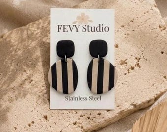 Puede incluir: Un par de pendientes a rayas negras y beige de FEVY Studio. Los pendientes tienen una parte superior cuadrada negra y una parte inferior ovalada beige con rayas negras verticales. La tarjeta dice "Stainless Steel".