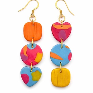 May include: Colorful dangle earrings with gold-tone hooks. Each earring features three vibrant shapes: a square, a circle, and a heart, in shades of orange, pink, blue, and yellow. The earrings are connected with small gold rings.