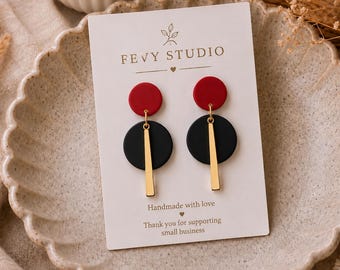 以下が含まれることがあります： FEVY STUDIOと書かれた白い台紙に固定された一対のぶら下がりピアス。各ピアスは上部に小さな赤い円、その下に大きな黒い円、そして黒い円の中心から垂れ下がる垂直の金色の金属棒で構成されています。