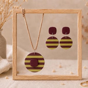 Puede incluir: Un conjunto de collar y pendientes exhibido en un marco de madera. La joyería presenta un diseño a rayas granate y amarillo. El collar tiene un colgante redondo con un acento cuadrado. Los pendientes son circulares con una parte superior cuadrada.