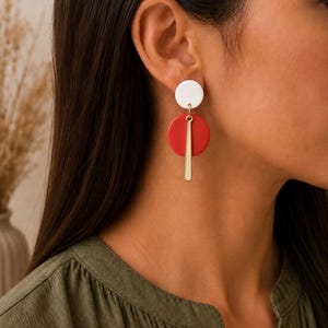 Puede incluir: Un pendiente colgante que presenta un peque&ntilde;o perno circular blanco conectado a un disco circular rojo m&aacute;s grande. Una barra de metal delgada y vertical de tono dorado cuelga del centro del disco rojo, creando un dise&ntilde;o geom&eacute;trico superpuesto.