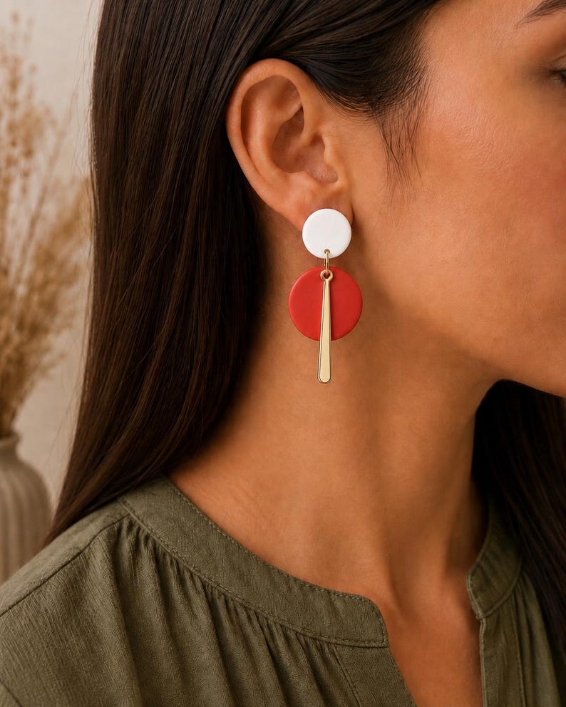 Puede incluir: Un pendiente colgante que presenta un peque&ntilde;o perno circular blanco conectado a un disco circular rojo m&aacute;s grande. Una barra de metal delgada y vertical de tono dorado cuelga del centro del disco rojo, creando un dise&ntilde;o geom&eacute;trico superpuesto.