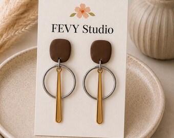 以下が含まれることがあります： FEVY Studioと書かれた白い台紙にセットされた一対のぶら下がりピアス。各ピアスは、ダークブラウンの四角いスタッド、シルバーカラーの金属リング、そしてリングの中央から吊り下げられた垂直のゴールドカラーの金属バーで構成されています。