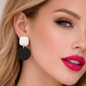 May include: Geometric earrings featuring a white square stud and a black circular drop. The earrings are shown on a person with blonde hair, green eyes, and bright pink lipstick.