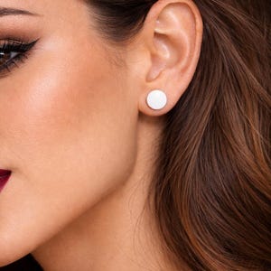 May include: A close-up of a person's ear with a white, circular stud earring. The earring is a simple, minimalist design. The person has brown hair and is wearing makeup.