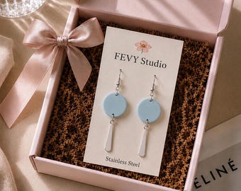 以下が含まれることがあります： FEVY Studioの台紙にセットされた、シルバーカラーの揺れるアクセントが付いたライトブルーの円形イヤリング。イヤリングは、同色のサテンリボンと茶色の紙製フィラーが入ったライトピンクのギフトボックスに収められています。