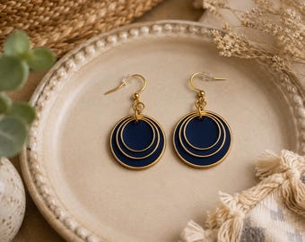 May include: A pair of drop earrings featuring a dark navy blue circular disc layered with two concentric gold-toned wire rings. The earrings are suspended from gold-toned fishhook ear wires, creating a tiered, geometric design with a smooth finish.