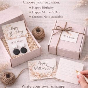 Puede incluir: Un set de regalo con dos cajas de cart&oacute;n rosa claro, una abierta con pendientes circulares blancos y negros sobre una tarjeta, y otra cerrada con cuerda. Incluye tarjetas con Happy Birthday y Happy Mother's Day, adem&aacute;s de una tarjeta en blanco.