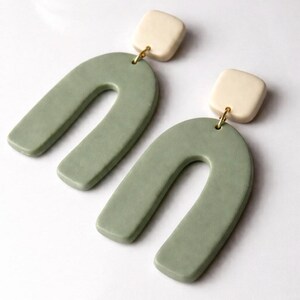 Peut inclure: Une paire de boucles d'oreilles modernes. Chaque boucle d'oreille est composée d'un carré crème relié à un pendentif vert sauge en forme de U, avec un connecteur doré. Les boucles d'oreilles sont présentées sur fond blanc.