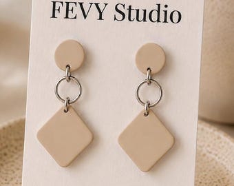 以下が含まれることがあります： FEVY StudioおよびStainless Steelと記載された白い台紙に固定されたベージュのイヤリング。各イヤリングは、小さなベージュの円形パーツと、銀色の金属リングで繋がれた大きなベージュのひし形ペンダントで構成されています。上部には小さなピンクの花のアイコンが印刷されています。