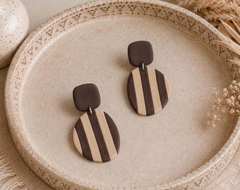 May include: A pair of dangle earrings featuring a dark brown square top piece connected to a larger, rounded bottom piece with vertical stripes in alternating shades of dark brown and beige. The earrings have a smooth, matte finish and a simple design.
