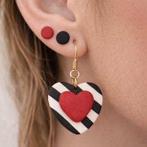 May include: A pair of earrings featuring a red heart on a black and white striped heart, with a gold-colored hook. The ear also has a red and a black stud earring.