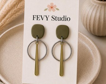以下が含まれることがあります： FEVY STUDIOと書かれた白い台紙にセットされた一対のぶら下がりピアス。各ピアスは、マットなオリーブグリーンの四角いスタッド、シルバーカラーの金属リング、そしてリングの中央から吊り下げられた垂直のゴールドカラーの金属バーで構成されています。