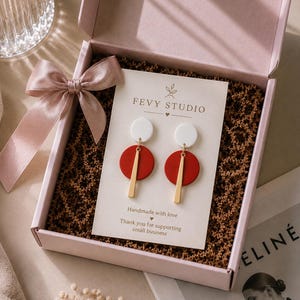 Puede incluir: Un par de pendientes colgantes con un c&iacute;rculo blanco arriba, un c&iacute;rculo rojo en el medio y una barra vertical dorada abajo, presentados en una tarjeta de FEVY STUDIO dentro de una caja de regalo rosa con cinta de sat&eacute;n y papel de relleno marr&oacute;n.