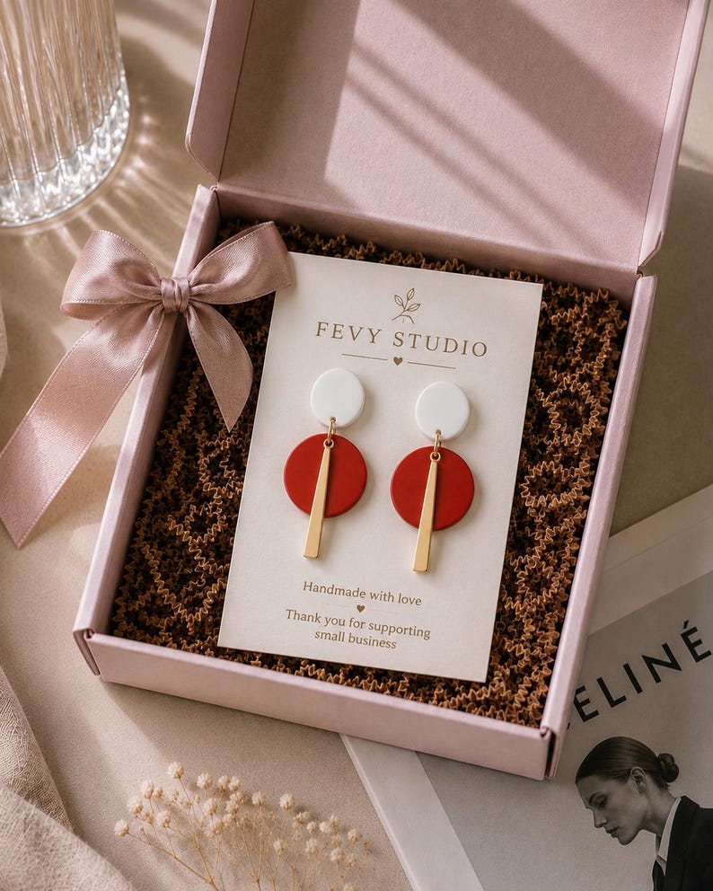 Puede incluir: Un par de pendientes colgantes con un c&iacute;rculo blanco arriba, un c&iacute;rculo rojo en el medio y una barra vertical dorada abajo, presentados en una tarjeta de FEVY STUDIO dentro de una caja de regalo rosa con cinta de sat&eacute;n y papel de relleno marr&oacute;n.