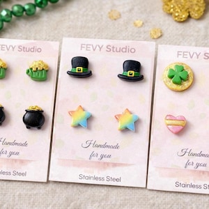 Puede incluir: Una colección de pendientes de acero inoxidable. Los pendientes presentan diseños temáticos del Día de San Patricio, incluyendo tazas verdes con oro, sombreros de copa negros, estrellas coloridas, tréboles, ollas de oro y corazones rosas. Las tarjetas dicen "Handmade for you."