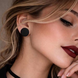 May include: A close-up of a person's face featuring a large, round, black earring. The person has blonde hair, dark eyeliner, and dark red lipstick. A black choker necklace is visible.
