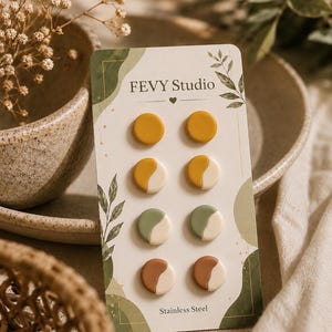 May include: A set of eight circular stud earrings on a FEVY Studio display card. The earrings feature solid mustard yellow, and two-toned designs in mustard yellow and cream, sage green and cream, and terracotta and cream. The card specifies Stainless Steel.