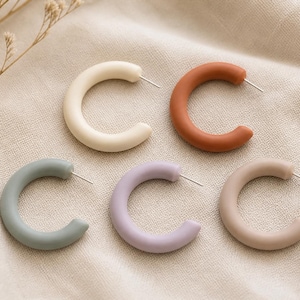 May include: Five colorful hoop earrings in cream, terracotta, sage green, lavender, and beige. The earrings are arranged on a neutral-toned fabric, showcasing their smooth, curved design and silver posts. A simple, modern jewelry set.