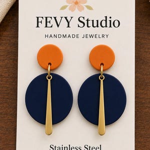 May include: A pair of dangle earrings on a white display card labeled FEVY STUDIO HANDMADE JEWELRY and Stainless Steel. Each earring features a small orange circle attached to a larger navy blue circle, accented by a vertical gold-tone metal bar.