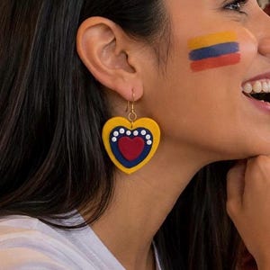 May include: Heart-shaped earrings with a yellow base, blue and red inner heart design, and white dot accents. The earrings are hanging from gold-colored hooks. The image shows a person wearing the earrings, with the Colombian flag painted on their cheek.
