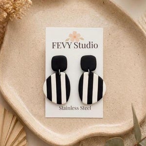 May include: Black and white striped earrings with a square top and oval bottom. The earrings are displayed on a white card with the text "FEVY Studio" and "Stainless Steel". The earrings are on a beige plate.