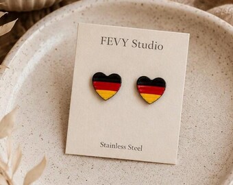 May include: A pair of heart-shaped stud earrings featuring the black, red, and gold horizontal stripes of the German flag. The earrings are attached to a cream-coloured display card printed with the text FEVY Studio and Stainless Steel.