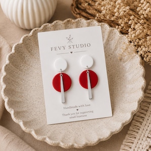 Puede incluir: Un par de pendientes en una tarjeta de presentaci&oacute;n blanca con la etiqueta FEVY STUDIO. Cada pendiente presenta un peque&ntilde;o c&iacute;rculo blanco en la parte superior, un c&iacute;rculo rojo m&aacute;s grande debajo y una fina barra vertical plateada colgando.