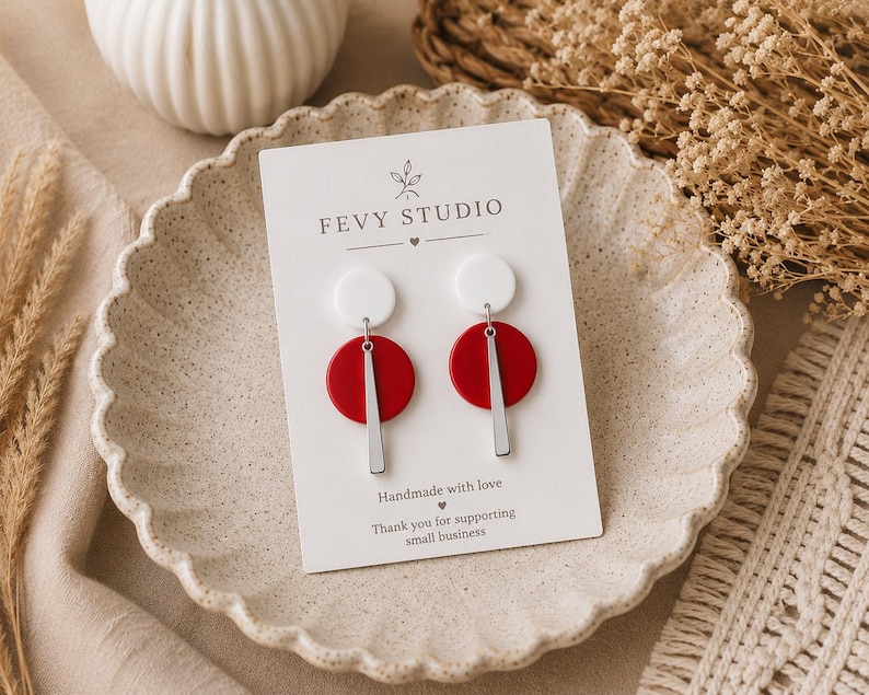 Puede incluir: Un par de pendientes en una tarjeta de presentaci&oacute;n blanca con la etiqueta FEVY STUDIO. Cada pendiente presenta un peque&ntilde;o c&iacute;rculo blanco en la parte superior, un c&iacute;rculo rojo m&aacute;s grande debajo y una fina barra vertical plateada colgando.