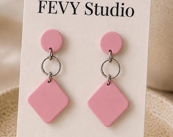 以下が含まれることがあります： FEVY Studioと書かれた白い台紙にセットされたピンク色のピアス。各ピアスは小さなピンクの円形パーツと、シルバーのリングで繋がれた大きなピンクのひし形ペンダントで構成されています。台紙には小さなピンクの花のグラフィックとStainless Steelの文字があります。