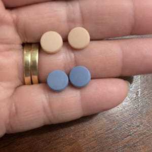 May include: Two pairs of small, round objects in a hand. One pair is a light tan color, and the other is a muted blue. The objects appear to be pills or small buttons.