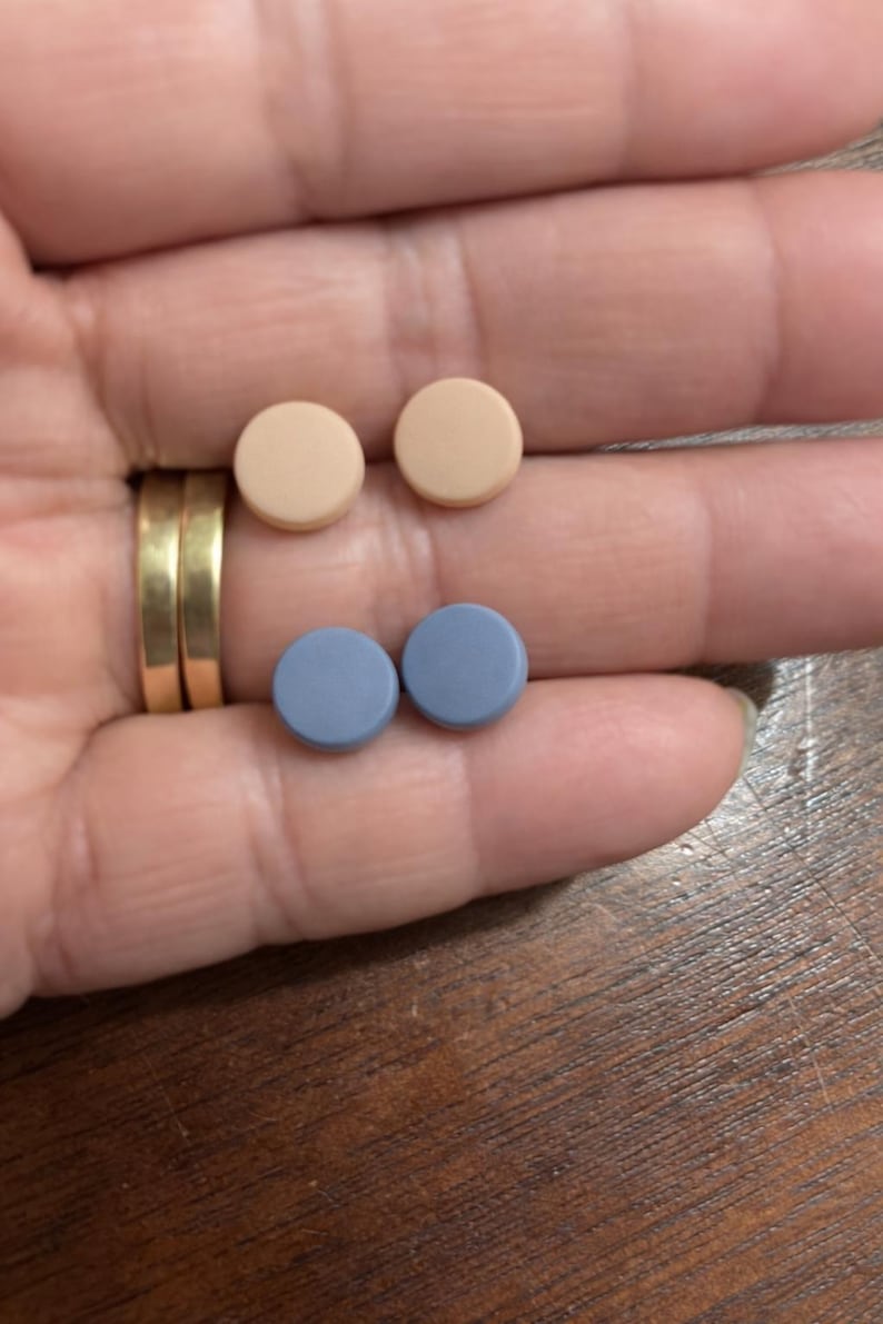 May include: Two pairs of small, round objects in a hand. One pair is a light tan color, and the other is a muted blue. The objects appear to be pills or small buttons.