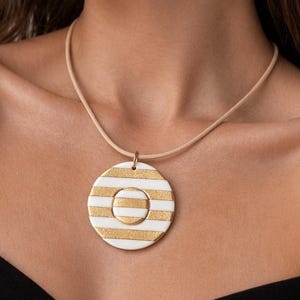 May include: A round pendant necklace with a cream-colored cord. The pendant is white with horizontal gold stripes and a smaller, matching center circle. The necklace is a stylish accessory.
