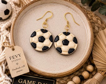 以下が含まれることがあります： 黒と白のサッカーボール柄の丸いドロップピアス。ピアスはゴールドトーンの金属製フックから吊り下げられています。各円形のチャームには、サッカーボールの古典的な五角形と六角形のデザインが施されています。