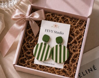 May include: A pair of dangle earrings featuring a small olive green square top connected to a larger circular pendant with vertical olive green and cream stripes. The earrings are displayed on a white card labelled FEVY Studio Stainless Steel.