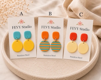 May include: Three pairs of dangle earrings on white display cards labelled FEVY Studio. Pair A has teal squares and yellow circles. Pair B has orange squares and orange-and-teal striped circles. Pair C has red squares and pale yellow circles.