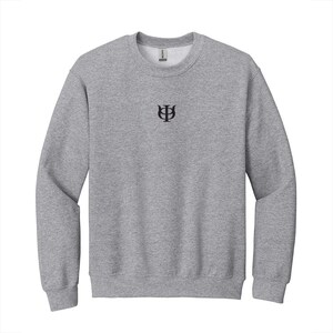May include: A heather gray crewneck sweatshirt with a small black embroidered symbol centered on the chest. The sweatshirt has a ribbed neckline, cuffs, and hem. The brand name is visible on the inside of the collar.