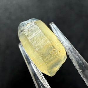 Puede incluir: Una piedra preciosa en bruto de color amarillo pálido con una estructura cristalina hexagonal alargada y un aspecto semitranslúcido. La piedra presenta estrías verticales naturales en sus lados y una textura superficial ligeramente desgastada.
