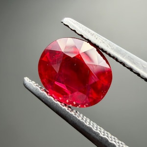 May include: A vibrant, oval-cut red gemstone held by a pair of silver-toned metal tweezers. The faceted stone displays a deep crimson hue with internal light reflections and sharp geometric cuts across its polished, translucent surface.