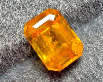 May include: A radiant, rectangular-cut gemstone with a vibrant orange hue. The gem's facets reflect light, creating a brilliant sparkle. The gemstone is set against a soft, gray background.