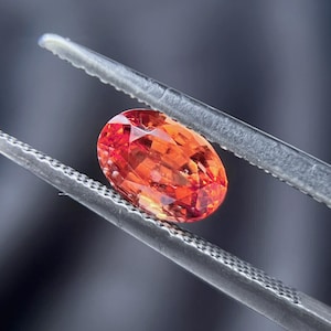 May include: An oval-cut, vibrant orange gemstone is held by a pair of silver tweezers. The gem's facets reflect light, showcasing its brilliance. The background is a blurred gradient of gray and blue.